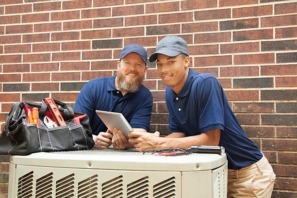 2 hvac technicians leaning on an outdoor unit and smiling