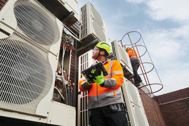 tech in hi-vis jacket looking up at large rooftop unit while on i pad
