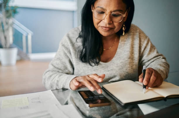 African woman writing in a journal creating a budget while using a mobile phone to calculate expenses. She carefully reviews financial details, focusing on managing her pension