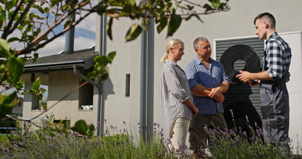 A technician is actively demonstrating the features of an external heat pump to a couple using a tablet. The scene captures a professional and informative interaction in a residential setting, enhanced by the natural surroundings of blooming lavender.