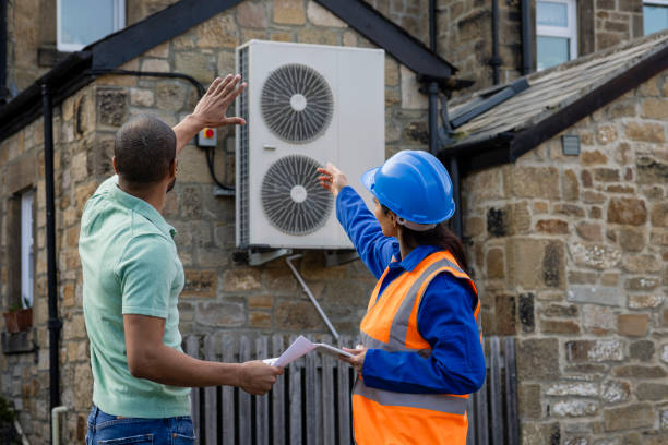 An air source heat pump on the side of a home being installed in the North East of England. The house is aiming to be sustainable. There is a construction worker with a hard hat and reflective jacket holding a digital tablet, they are both pointing to the heat pump.