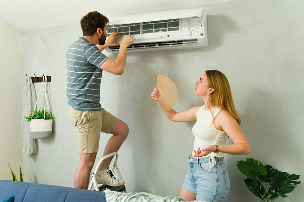 Caucasian man standing on a ladder installing or repairing a mini split ac unit while a woman fans herself, waiting for cool air