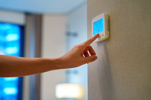 A woman using a smart home thermostat to adjust room temperature in a modern apartment, illustrating home automation, energy efficiency, and technology in daily life.