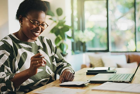 Happy, black woman and finance with laptop for payroll, tax return or budget planning at home. Female person, accounting or smile with calculator, documents or bills for expenses, deductions or audit